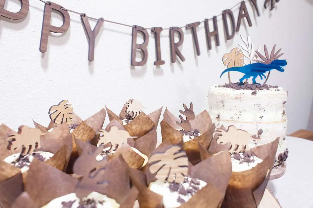 Dinosaur party pack, teal T-REX cake topper displayed on cupcakes and a cake, with a happy birthday banner hanging on the wall.