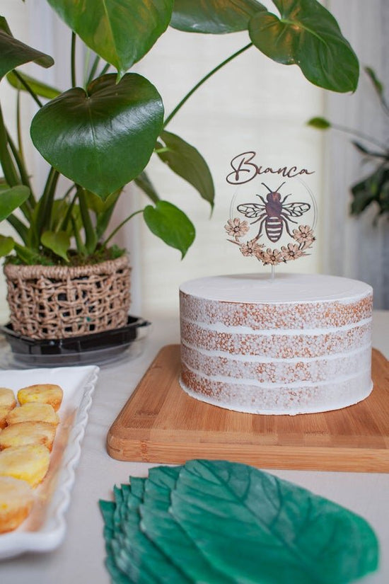 Custom bee cake topper, bee topper with name on a cake next to party decor and green plants.