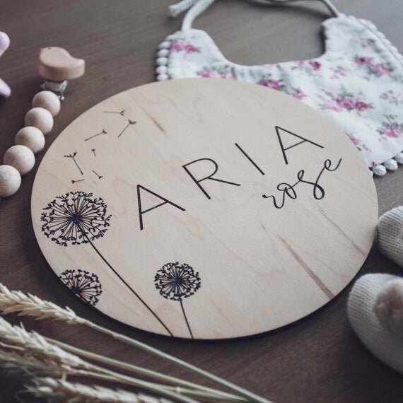 Dandelion engraved birth announcement plate next to baby items and botanical plant.