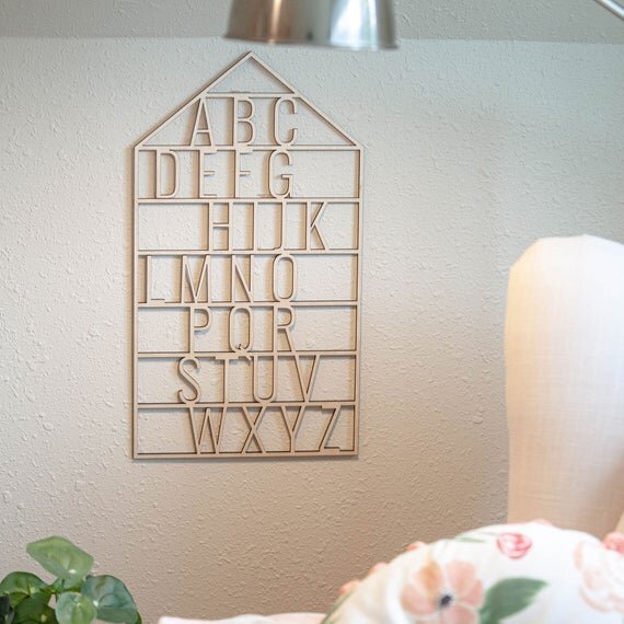 wooden alphabet house nursery decor next to a white chair, lamp, and a green plant