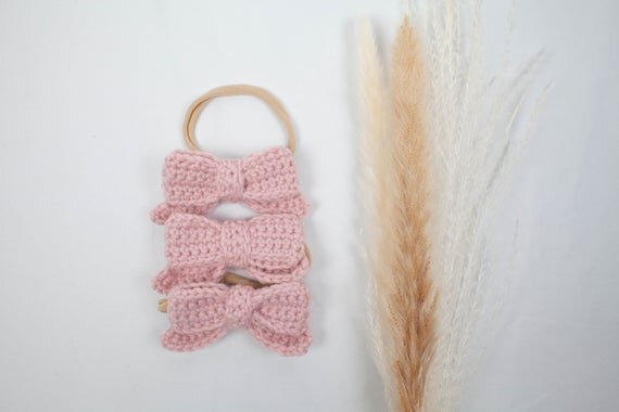 Blush color crochet hair bows with head bands displayed next to botanical plant.