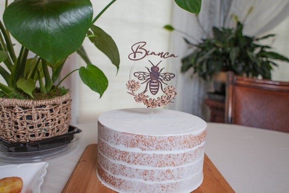 Load image into Gallery viewer, Custom bee cake topper, bee topper with name on a white cake with green plants in the background.
