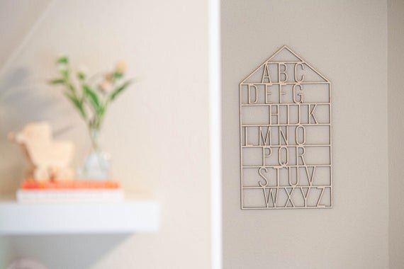 wooden alphabet house nursery decor on a white wall next to a shelf that has books and a plant in a vase