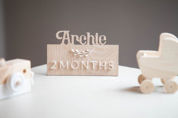 Custom wooden baby milestones, 3D milestone plaques with name next to wooden camera toy and wooden toy stroller.