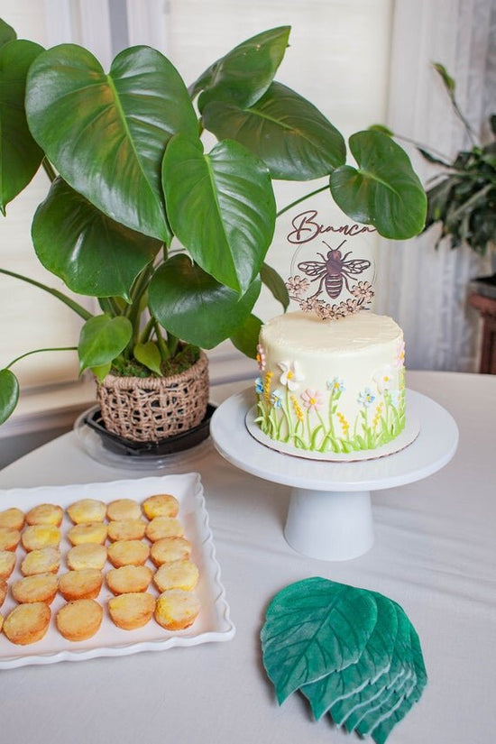Custom bee cake topper, bee topper with name on a cake next to party decor and green plants.