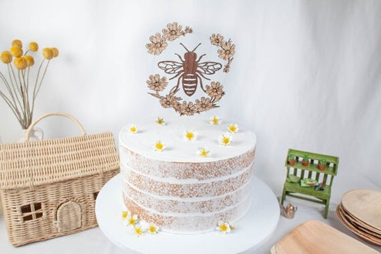 Bee Cake Topper with Flowers, birthday bee cake topper on a white cake, next to plates and botanical decor.