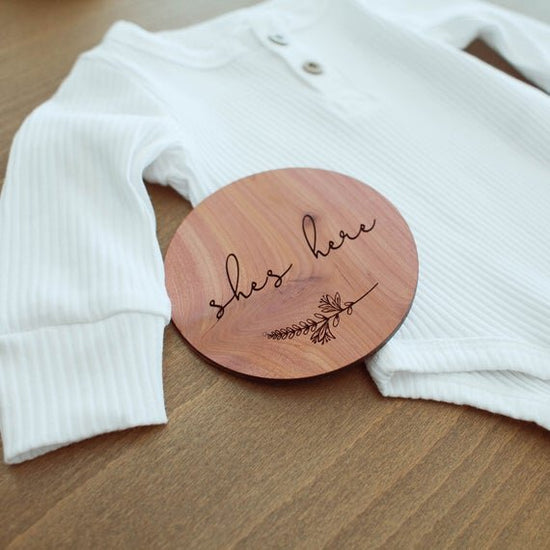 Load image into Gallery viewer, She&amp;#39;s here red cedar wood birth announcement circle next to baby white long sleeve onesie

