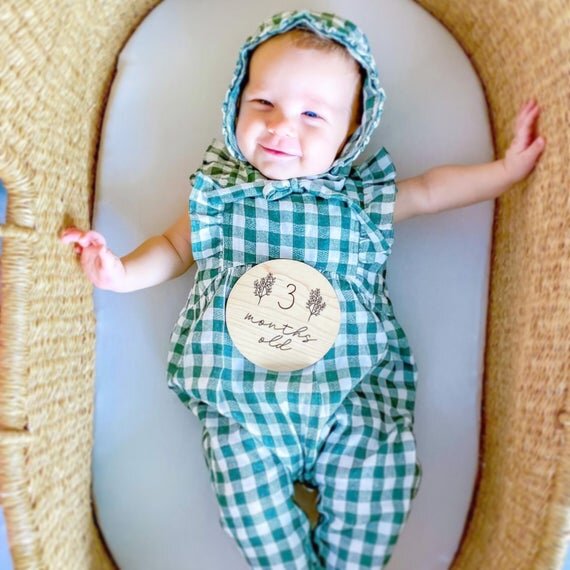Botanical wooden baby milestone circle on top of 3 month baby in a blue onesie inside bassinet.