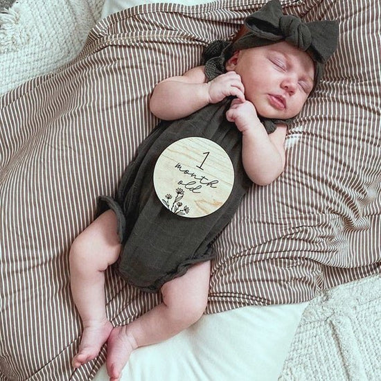 Botanical wooden baby milestone circle on top of 1 month baby in a black onesie laying on a bed.