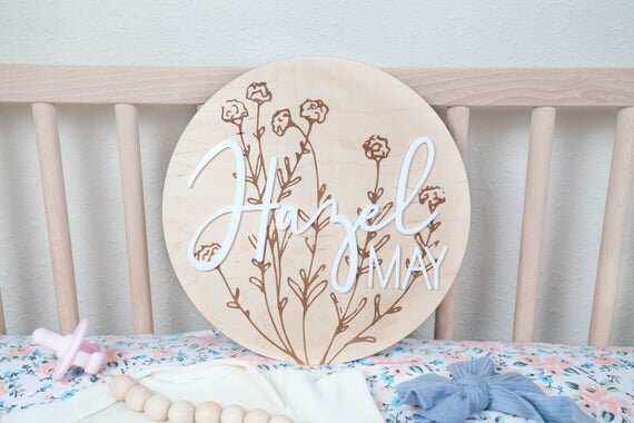 baby girl nursery name sign with botanical details inside wooden crib next to pink pacifier, white onesie, and blue bow