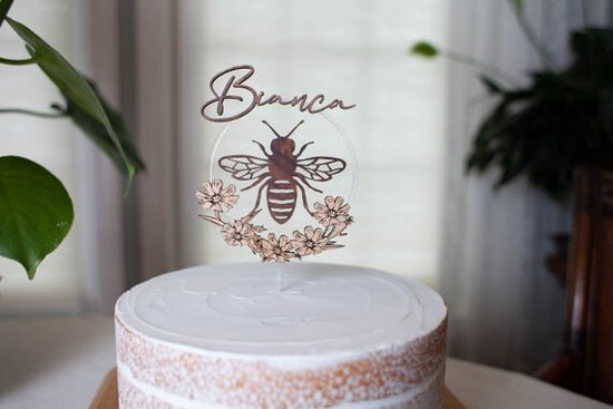 Custom bee cake topper, bee topper with name on a cake next to green plants.
