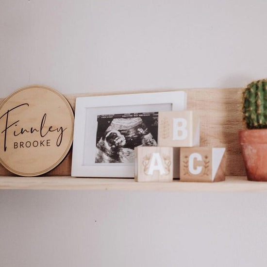 Baby name sign, welcoming home baby name sign, minimalistic birth announcement, on wooden shelf next to baby ultrasound, alphabet bricks, and a cactus.