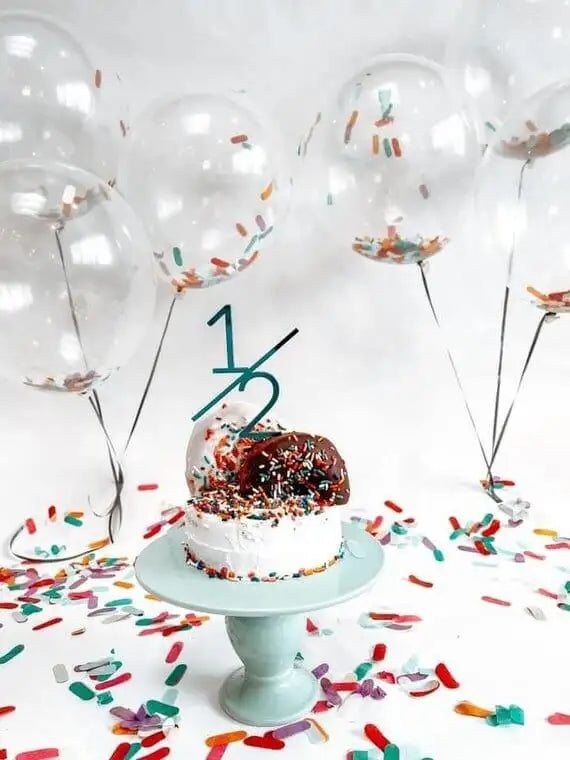 1/2 birthday cake topper on a sprinkled cake on top of a blue cake stand with balloons filled with confetti