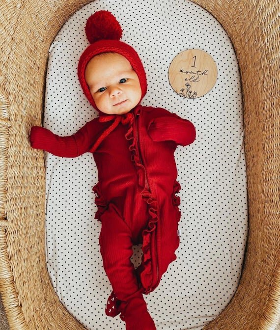 Botanical wooden baby milestone circle on top of 1 month baby in a red onesie inside bassinet.
