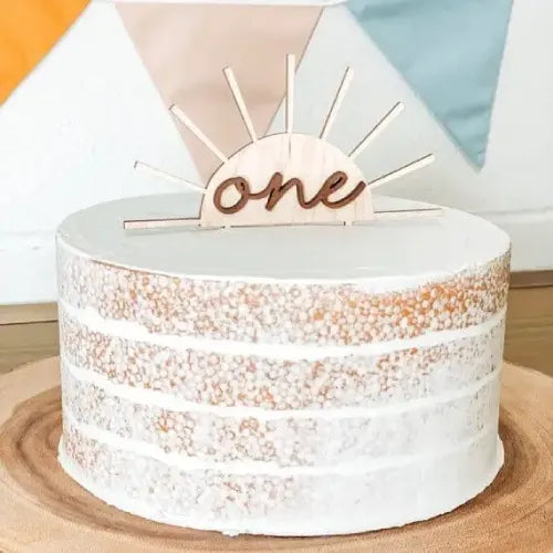 Boho sun cake topper, boho one year sun cake topper on white cake, on top of wooden platter next to hanging colorful banner.