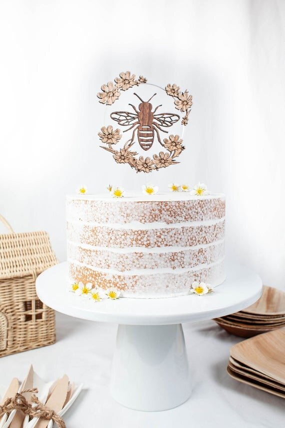 Bee Cake Topper with Flowers, birthday bee cake topper on a white cake next to plates and utensils.
