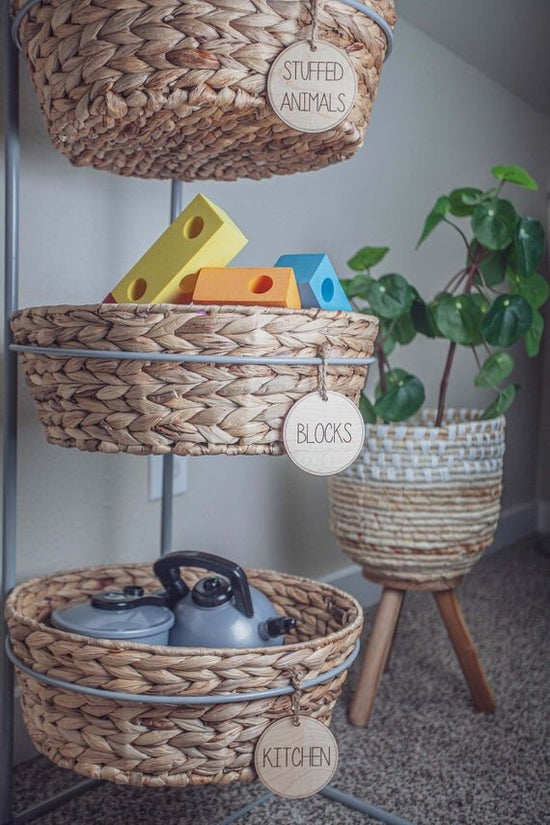 3" circular wooden toy storage tags on a 3 tier wicker baskets filled with toys and green plant in the background