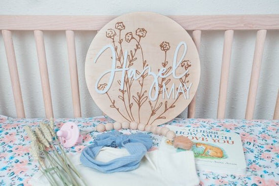 baby girl nursery name sign with botanical details inside wooden crib next to pink pacifier, white onesie, a blue bow, a book, and botanical plants
