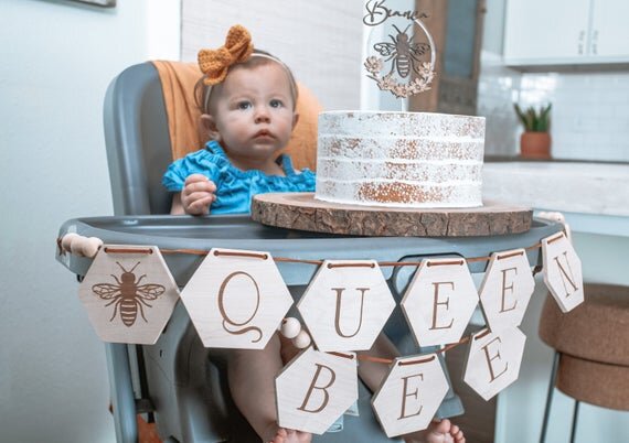 Custom bee cake topper, bee topper with name on a cake on top of a high chair with baby sitting on it, with queen bee banner hanging off the chair.