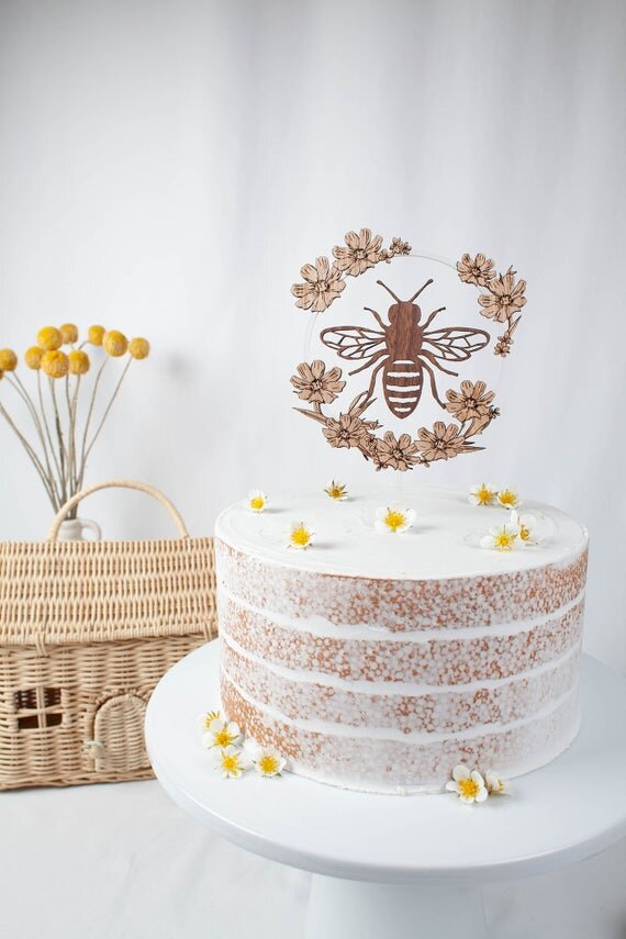Bee Cake Topper with Flowers, birthday bee cake topper on a white cake next to botanical decor.
