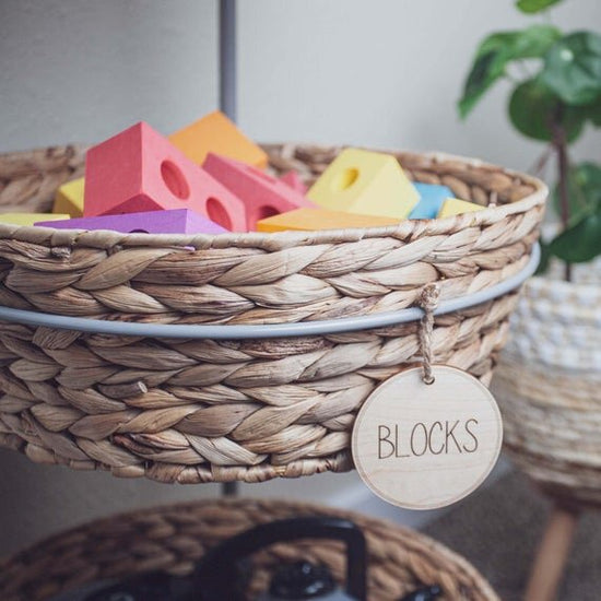 3" circular wooden toy storage tags on a wicker basket filled with toy blocks and green plant in the background