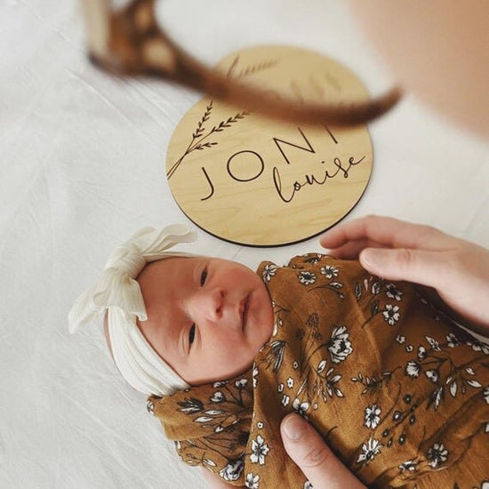 Botanical engraved wooden baby sign next to swaddled newborn baby being observed from above by the mother.