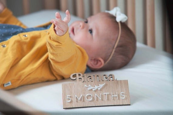 Custom wooden baby milestones, 3D milestone plaques with name next to laying down baby in wooden crib.