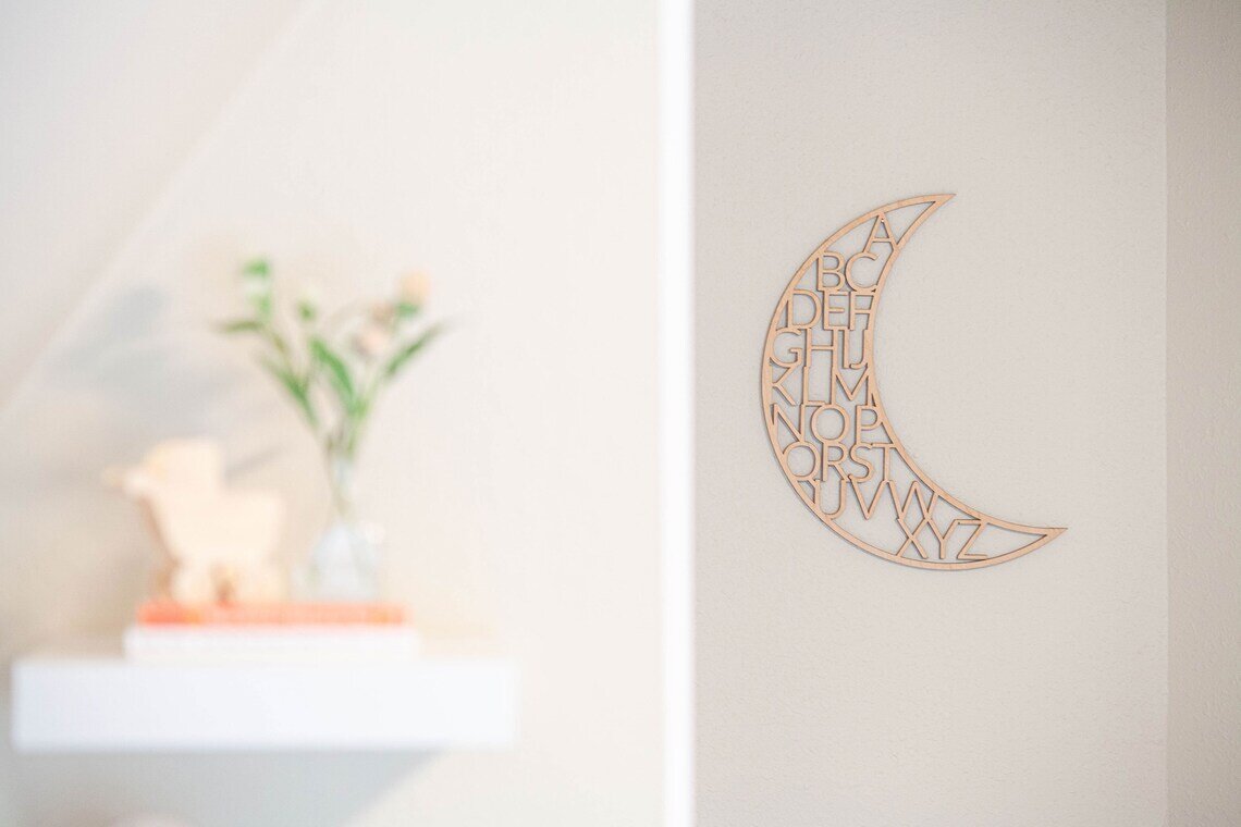 wooden alphabet moon nursery decor on a white wall next to a shelf that has books and a plant in a vase